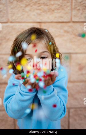 Fille soufflant confetti colorés devant le mur Banque D'Images