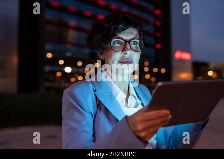 Belle femme d'affaires mûre portant des lunettes tenant une tablette numérique la nuit Banque D'Images
