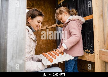 Mère tenant le contenant pendant que les filles arrangeant les oeufs dans la ferme Banque D'Images