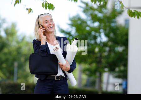 Femme d'affaires souriante tenant des plans tout en parlant sur un smartphone Banque D'Images