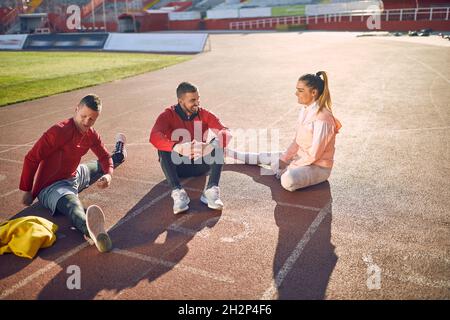 jeune adulte caucasien beardy gars assis sur la piste d'athlète avec le sourire couple à qui parle. concept entraîneur personnel Banque D'Images