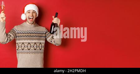 Concept de vacances d'hiver, noël et célébration. Un gars adorable et enthousiaste en chandail et chapeau de père noël, en appréciant la fête du nouvel an, en levant le verre et Banque D'Images
