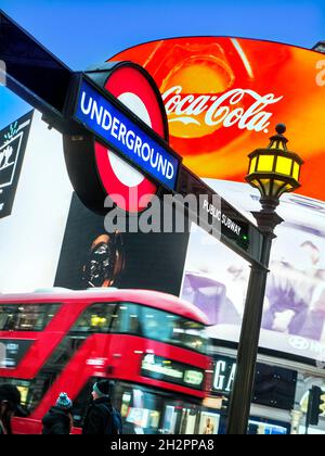 Londres Piccadilly Circus avec panneau de métro traditionnel souterrain et panneau d'affichage numérique Coca Cola néon allumé derrière et rouge flou hybride à faibles émissions London bus au crépuscule Piccadilly Londres UK Banque D'Images