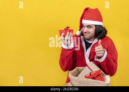 Un homme enthousiaste habillé comme le Père Noël montre un petit cadeau rouge pris dans un grand sac rempli de cadeaux et exprime son apprecciation avec le ventre vers le haut Banque D'Images