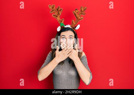 Jeune fille hispanique portant le chapeau de noël cerf choqué couvrant la bouche avec des mains pour erreur. Concept secret. Banque D'Images