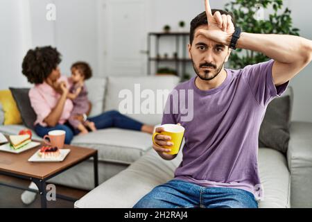 Père hispanique de famille interraciale boire une tasse de café rendant amusant des gens avec les doigts sur le front faisant le geste perdant mocking et insultant. Banque D'Images
