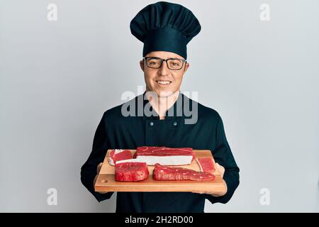 Beau jeune homme portant un uniforme de chef, portant un plateau de maintien avec de la viande crue, souriant avec un sourire heureux et frais sur le visage. Montrant les dents. Banque D'Images