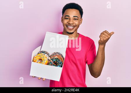 Jeune homme afro-américain tenant une boîte avec des beignets colorés savoureux pointant du pouce vers le côté souriant heureux avec la bouche ouverte Banque D'Images