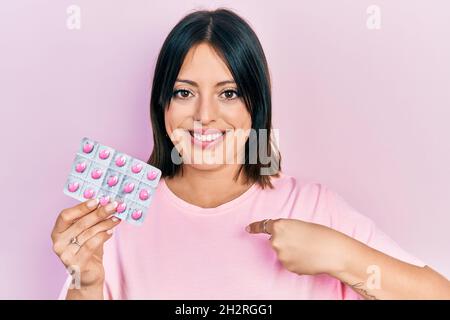 Jeune femme hispanique tenant des pilules pointant du doigt vers un individu souriant heureux et fier Banque D'Images