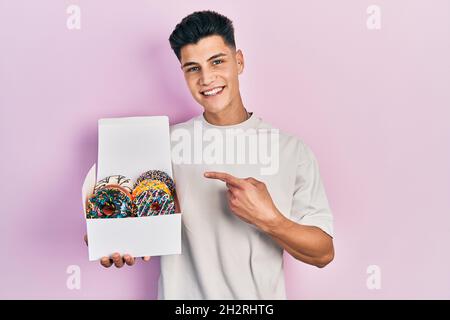 Jeune homme hispanique tenant une boîte de beignets colorés et savoureux souriant, souriant et souriant, avec la main et le doigt Banque D'Images