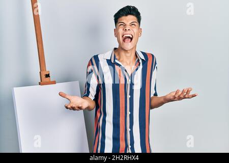 Jeune hispanique se tenant près de la toile vide fou et fou criant et hurlant avec l'expression agressive et les bras levés. Notion de frustration. Banque D'Images