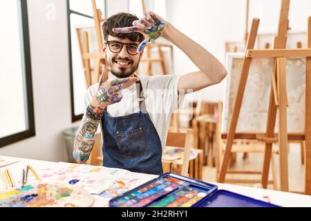 Homme hispanique à la barbe dans un studio d'art souriant faisant cadre avec les mains et les doigts avec le visage heureux. Créativité et concept de photographie. Banque D'Images