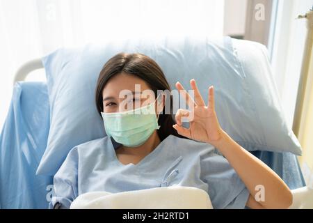 Une patiente asiatique heureuse porte un masque, repose sur le lit et affiche un symbole ok pour montrer la confiance dans le traitement.Concept de croire en traitement et Banque D'Images