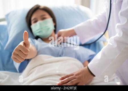 Une patiente asiatique heureuse porte un masque, se repose sur le lit et lève le pouce.Lorsqu'un médecin utilise un stéthoscope pour écouter les poumons.Concept de Banque D'Images
