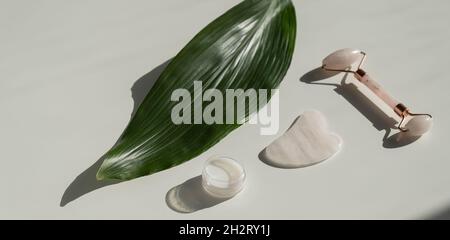 Grattoir de gouache rose, rouleau, pot de crème de feuille verte sur une table blanche. Équipement pour l'auto-massage et les soins de la peau pour le visage et le cou Banque D'Images