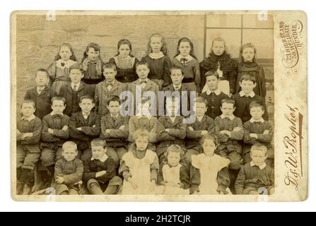 Original, teinte sépia carte victorienne d'armoire d'écoliers victoriens, jeunes et jeunes garçons et filles, visages sérieux, posant pour un portrait de groupe à l'extérieur dans la cour de l'école. Beaucoup de modes merveilleuses du début à la fin des années 1890 - robes smock, et les styles de manches bouffées de 1893-98, colliers en dentelle et certains des garçons portent des noeuds papillon. Assis les bras pliés. Du studio de D. & W. Prophet, Dundee, Écosse, Royaume-Uni photo prise vers 1896 Banque D'Images