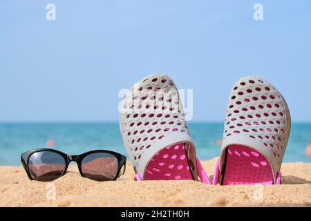 Gros plan sur les chaussures Clogs et les lunettes de soleil noires protectrices sur la plage de sable au bord de la mer tropicale par temps chaud et ensoleillé. Concept de vacances d'été. Banque D'Images