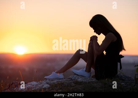 Jolie femme triste en robe d'été courte noire assise sur un rocher pensant à l'extérieur au coucher du soleil. Femme à la mode contemplant une soirée chaude dans la nature Banque D'Images