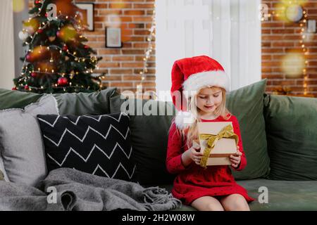 Petite fille souriante en robe rouge festive tenant cadeau de Noël ou de nouvel an.Enfant caucasien vêtu d'un chapeau de père Noël ou d'une casquette Banque D'Images