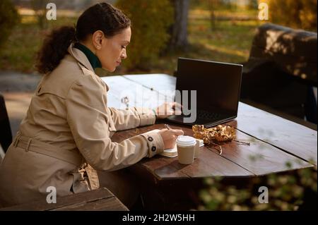 Femme d'affaires confiante, sous un manteau beige décontracté, assise sur un banc en bois dans un café et travaillant à distance, prend des notes sur un ordinateur portable et les types sur un clavier d'ordinateur portable Banque D'Images