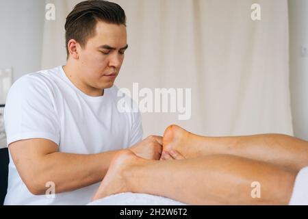 Gros plan court de masseuse professionnelle de sexe masculin avec des mains fortes massant les pieds talons de l'athlète musclé homme après l'entraînement physique Banque D'Images