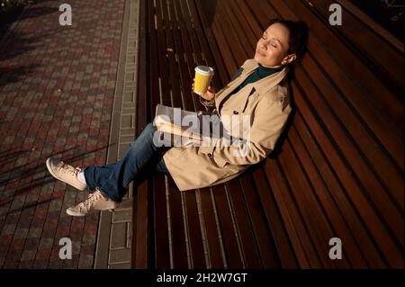 Belle et paisible jeune femme est assise sur un banc en bois, penchée sur son dos dans un parc, tient une tasse en papier jetable avec boisson chaude et un livre, se reposant Banque D'Images