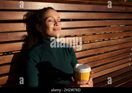 Gros plan d'une charmante jeune femme paisible assise sur un banc en bois, penchée sur son dos dans un parc, tient une tasse en papier à emporter avec un restin à boisson chaude Banque D'Images