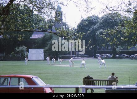 Un match de cricket auquel participent des membres du club de cricket de Kew, à Kew Green, à l'ouest de Londres, au début des années 1970.Les spectateurs regardent le match depuis un banc de parc et l'église Sainte-Anne se trouve au-delà du terrain de cricket. Banque D'Images