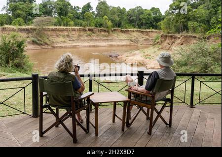 Couple caucasien assis sur la zone d'observation au camp de Governors observant les hippopotames près de la rivière Mara à distance, Masai Mara, Kenya Banque D'Images