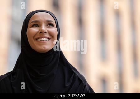 Gros plan de jeune femme islamique souriante dans le hijab noir debout à l'extérieur Banque D'Images