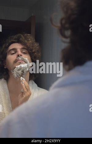 Un jeune homme en peignoir applique de la mousse de rasage sur le visage dans la salle de bains de l'appartement.Concept de spa Beauty Home. Banque D'Images