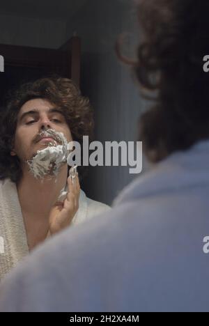 Un jeune homme en peignoir applique de la mousse de rasage sur le visage dans la salle de bains de l'appartement.Concept de spa Beauty Home. Banque D'Images