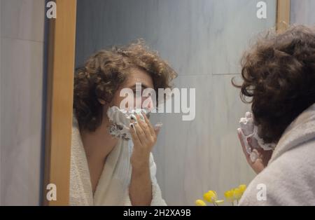 Un jeune homme en peignoir applique de la mousse de rasage sur le visage dans la salle de bains de l'appartement.Concept de spa Beauty Home. Banque D'Images