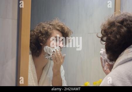 Un jeune homme en peignoir applique de la mousse de rasage sur le visage dans la salle de bains de l'appartement.Concept de spa Beauty Home. Banque D'Images