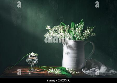 Classique encore la vie avec le beau blanc Lily de la vallée bouquet en pot blanc et un verre d'eau en rayon de lumière.Photographie d'art. Banque D'Images