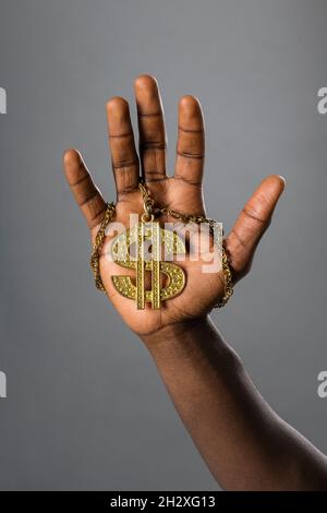 Homme noir tenant un symbole de Dollar d'or décoratif sur un collier l'affichant sur la paume de sa main avec la chaîne enroulée autour de ses doigts Banque D'Images