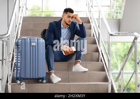 Un gars du Moyen-Orient contrarié assis sur les escaliers du terminal de l'aéroport avec ses bagages