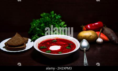 Gros plan d'une assiette de borsch russe rouge avec du pain, des légumes et des légumes verts en arrière-plan. Banque D'Images