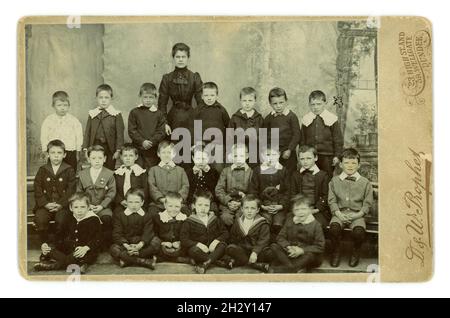 Carte originale de cabinet victorien teinte sépia d'écoliers victoriens juniors, classe de garçons, posant pour un portrait de studio en plein air, avec leur professeur à l'apparence stricte. Il y a une toile de fond en bambou d'influence japonaise. Tous les enfants sont des garçons, le professeur à la recherche sévère a des manches bouffées et une taille minuscule, sélection intéressante de cols et de vêtements pour garçons, de nœuds papillon et de costumes de marin, du studio de D. & W. Prophet, Dundee, Écosse, Royaume-Uni vers 1898 Banque D'Images