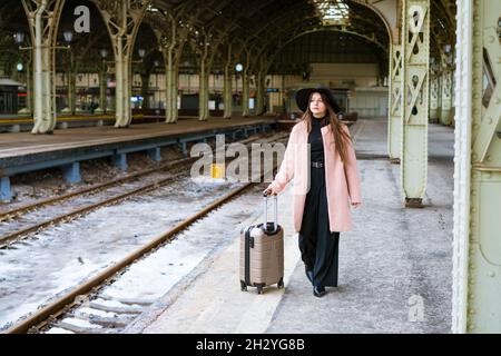 Une jeune femme en manteau marche avec une valise le long de la plate-forme vide de la gare.Caucasien fille touriste.Concept de voyage seul Banque D'Images