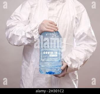 un homme en costume de protection et gants tient une grande bouteille en plastique avec l'inscription covid 19 vaccin. Banque D'Images