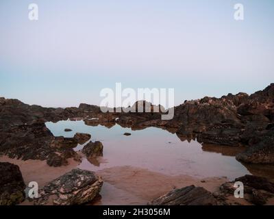 Couleurs du crépuscule, scène côtière. Rose pastel et ciel bleu reflété dans l'eau encore brillante des piscines de roche en dessous sur la plage, coucher de soleil Banque D'Images