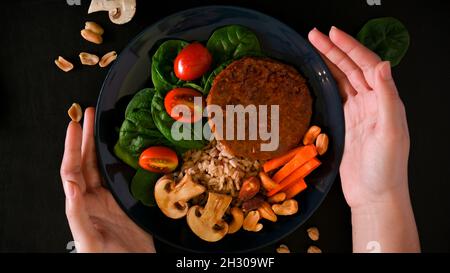 Vue de dessus gros plan des mains de femmes tenant une assiette de légumes à base de plantes. Cutlet végétalien sans viande, riz brun et divers légumes frais en b Banque D'Images