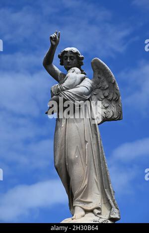 Une sculpture en pierre d'un ange, un de ses doigts cassés, tenant un bébé, avec ciel bleu et nuages blancs clairs en arrière-plan Banque D'Images