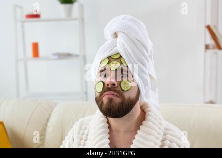 Homme barbu tenant des tranches de concombre sur le visage. Spa à la maison, soins du corps et de la peau pour homme concept. Banque D'Images