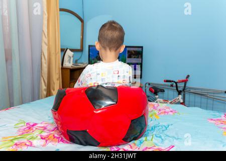 Un petit garçon est assis dans un fauteuil à sac de beanbag souple pour enfants sous forme de football rouge sur le lit et regardant la télévision Banque D'Images