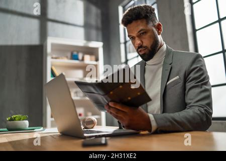 Jeune homme d'affaires afro-américain avec journal travaillant sur ordinateur portable à l'intérieur dans le bureau Banque D'Images