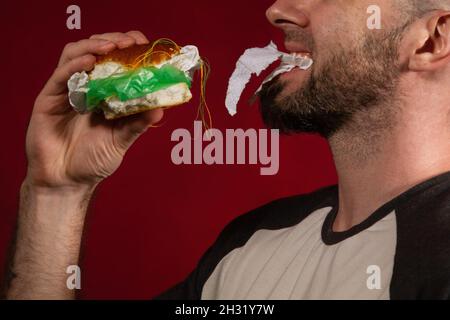 Le concept de restauration rapide et d'écologie.Un homme chauve avec une barbe, mangeant heureusement un hamburger bourré de déchets.Arrière-plan Bourgogne.Gros plan. Banque D'Images