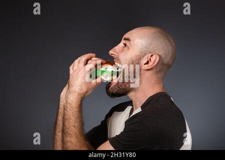 Le concept de restauration rapide et d'écologie.Un homme chauve avec une barbe, mangeant heureusement un hamburger bourré de déchets.Une mâchoire grotesquely élargie.Dos noir Banque D'Images