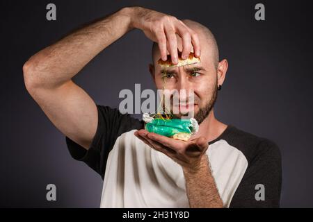 Le concept de restauration rapide et d'écologie.Un homme chauve avec une barbe regarde la farce d'un hamburger désassemblé avec horreur.Arrière-plan sombre. Banque D'Images